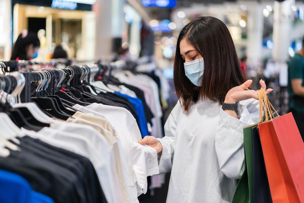 
                        Frau mit Hygienemaske und Einkaufstaschen schaut sich Kleider in einem Modegeschäft an
                                              